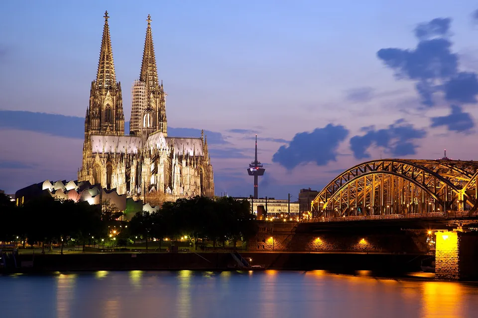 Beleuchteter Kölner Dom und Hohenzollernbrücke mit Spiegelungen im Rhein zur blauen Stunde.