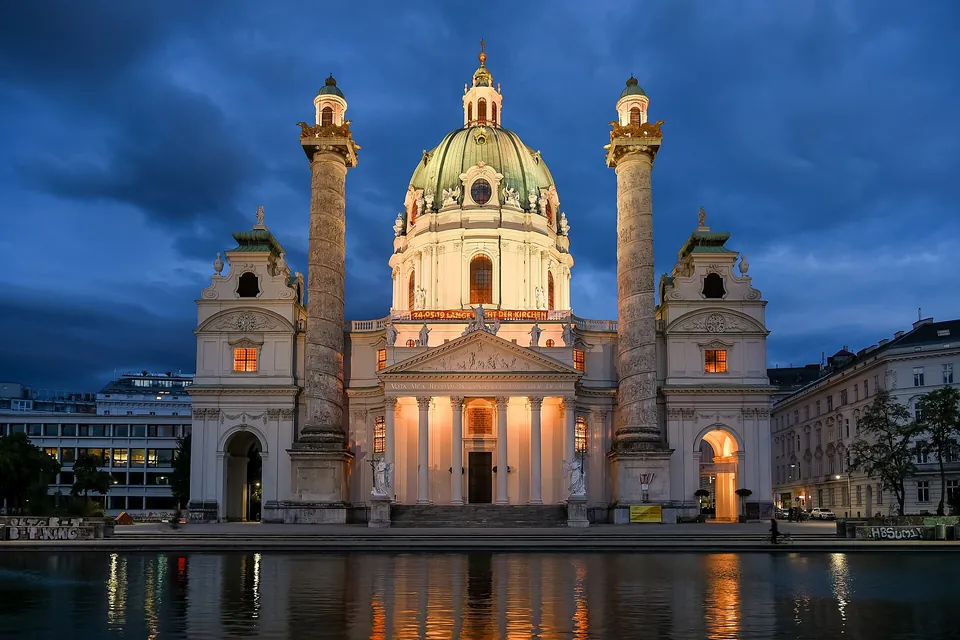 Wiener Karlskirche mit beleuchteter Kuppel und Spiegelungen im Wasser zur blauen Stunde.